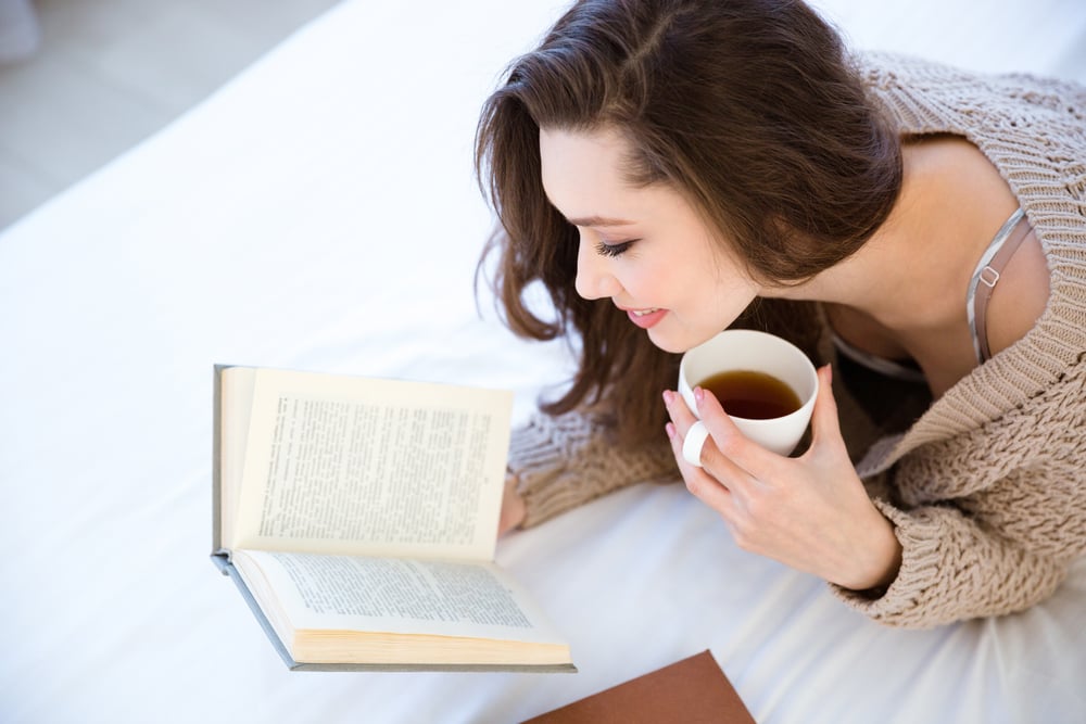 Cute lovely young woman reading book and drinking coffee on bed