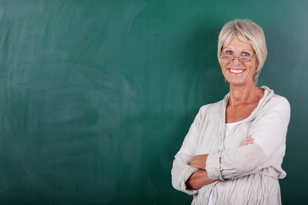 Portrait of happy senior female teacher with arms crossed standing against chalkboard